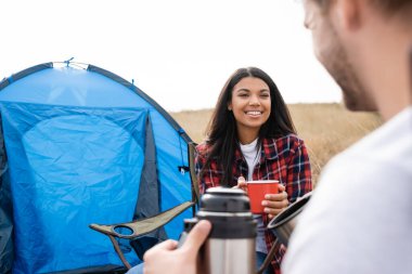 Cheerful african american woman holding cup near boyfriend with thermos on blurred foreground and tent outdoors  clipart