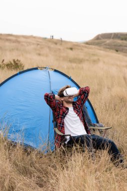 VR kulaklıklı gülen adam çimenli çayırdaki çadırın yanındaki sandalyede oturuyor. 