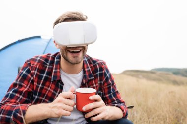 VR kulaklıklı gülümseyen adam çadır yanında fincanla çayırda bulanık arka planda. 