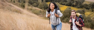 Cheerful multicultural hikers walking with backpacks on field, banner  clipart