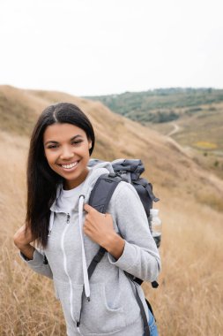 Cheerful african american hiker with backpack looking at camera with blurred landscape at background  clipart
