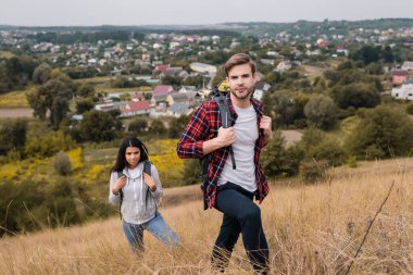 Couple of multiethnic travelers with backpacks looking at camera on grassy hill  clipart