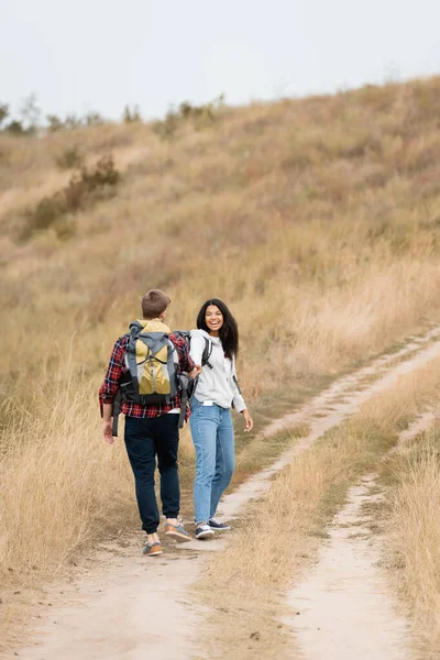Smiling African American Woman Looking Boyfriend Backpack While Walking Path — Stock Photo, Image