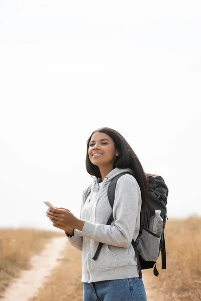 Mujer Afroamericana Sonriente Con Mochila Sosteniendo Teléfono Inteligente Con Camino — Foto de Stock