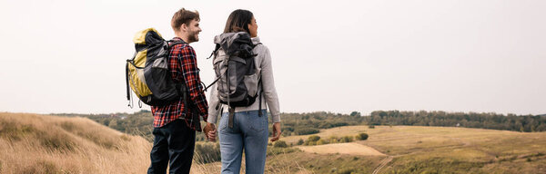 Smiling interracial couple with backpacks holding hands with grassy hills on blurred background, banner 