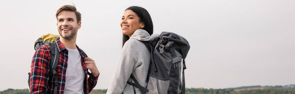 Smiling multiethnic hikers with backpacks looking away outdoors, banner 