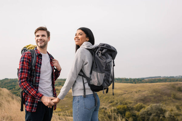 Smiling multiethnic couple with backpacks holding hands with hills on blurred background 
