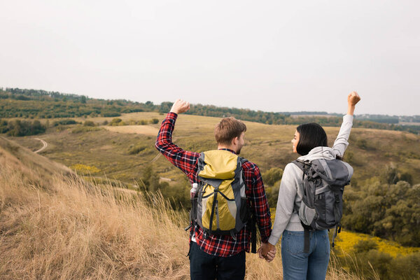Улыбающаяся африканская американка с рюкзаком, показывающая да жест и держащая за руку парня с холмами на размытом фоне 