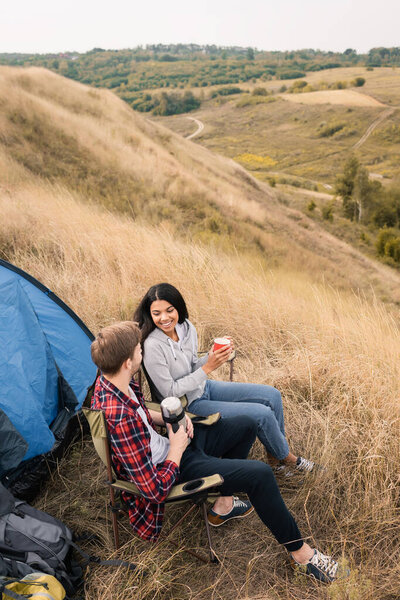 Smiling interracial couple holding cup and thermos near tent during camping in field  