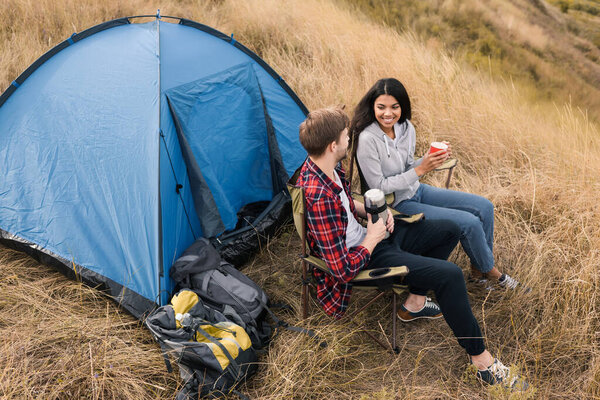 African american woman holding cup near boyfriend with thermos and tent on grassy lawn 