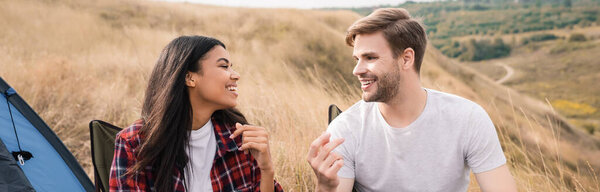 Cheerful man talking to african american girlfriend on chairs during camping on lawn, banner 