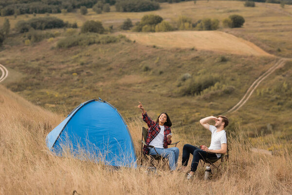 Smiling african american woman holding smartphone and pointing with finger near boyfriend with cup and tent on lawn 