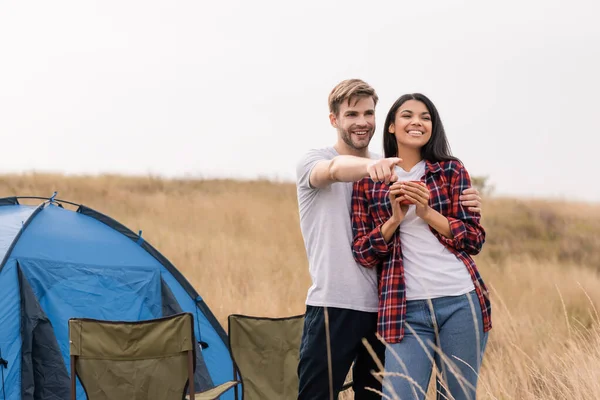 Smiling Man Embracing African American Girlfriend Cup Pointing Finger Camping — Stock Photo, Image