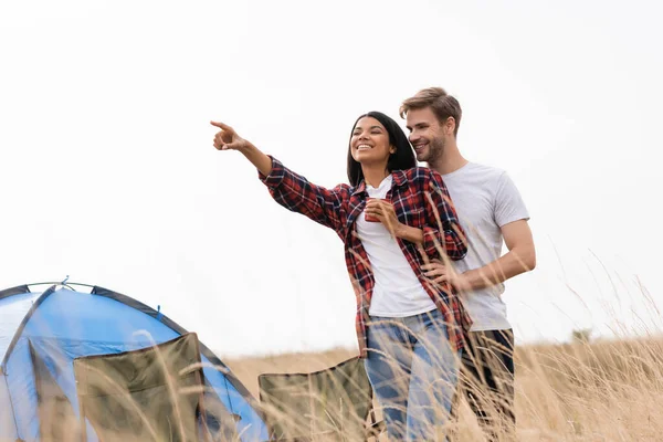 Hombre Sonriente Abrazando Novia Afroamericana Con Taza Apuntando Con Dedo — Foto de Stock