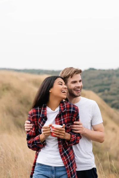 Hombre Alegre Abrazando Mujer Afroamericana Con Taza Campo Herboso Durante — Foto de Stock