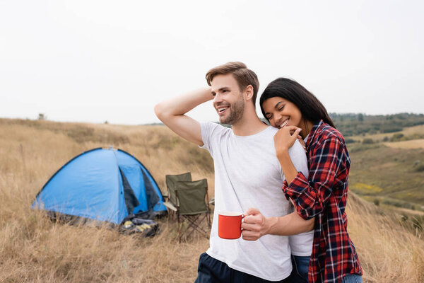 African american woman with closed eyes hugging boyfriend with cup near tent on blurred foreground 