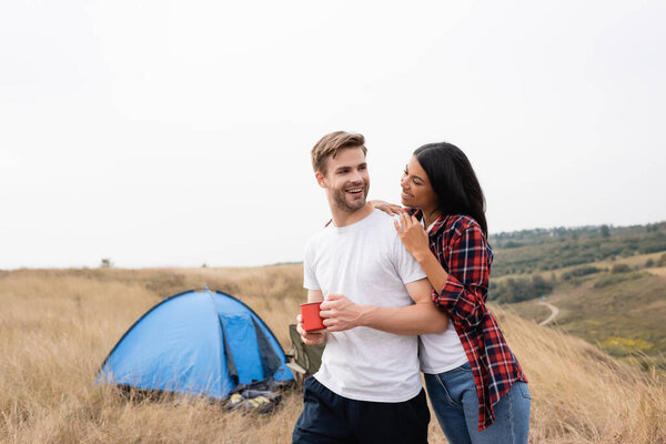 Smiling african american woman hugging boyfriend with cup near tent on blurred background outdoors 