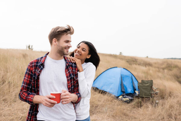 Smiling man holding cup near african american girlfriend during camping in field 