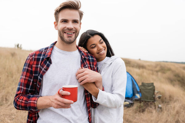 Smiling african american woman holding hand of boyfriend with cup during camping in field 