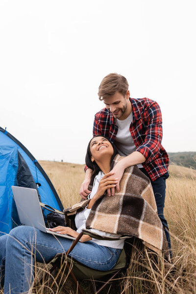 Multiethnic couple with blanket and laptop smiling at each other near tent on meadow 