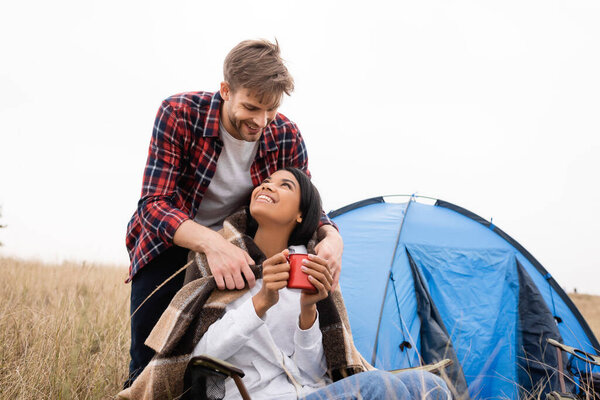 Smiling man putting blanket on smiling african american girlfriend with cup near tent