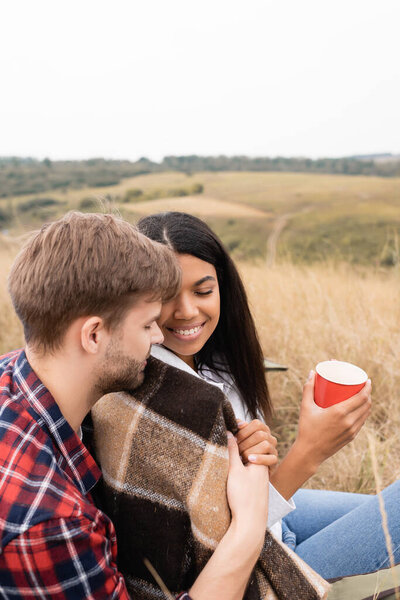 Man hugging smiling african american woman in blanket with cup on grassy meadow 