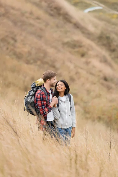 Smiling Multiethnic Couple Backpacks Holding Hands Grassy Hill — Stock Photo, Image