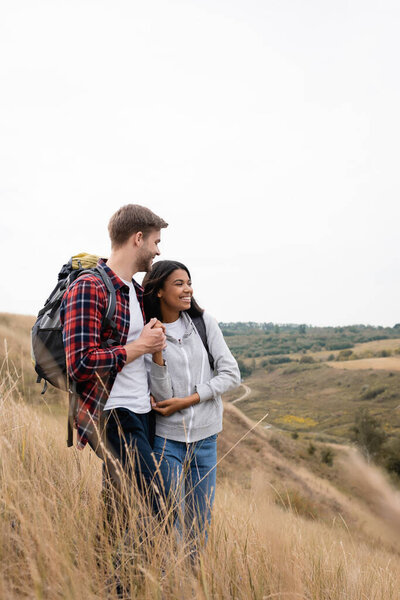 Smiling interracial couple of tourists holding hands while standing in grass on hill 