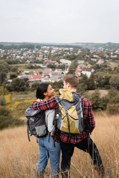 Back view of tourist with backpack hugging smiling african american girlfriend on grassy hill