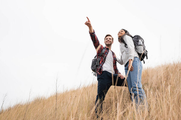 Low angle view of smiling man with backpack pointing with finger away near african american woman while standing on grassy hill with sky at background 