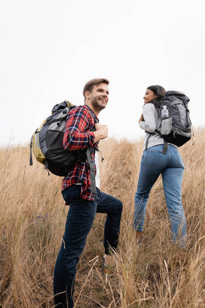Smiling man holding backpack while standing near african american girlfriend on hill with grass 