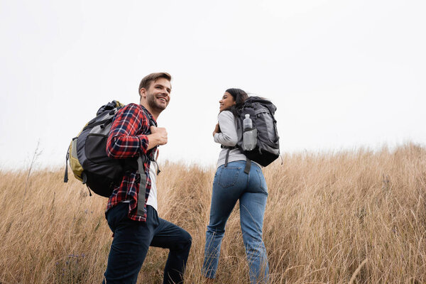 Young hiker smiling near african american girlfriend walking on grassy hill 