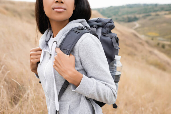 Cropped view of african american woman with backpack standing on hill with blurred landscape at background 