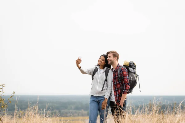 Sorrindo Casal Interracial Com Mochilas Falando Selfie Com Smartphone Gramado — Fotografia de Stock