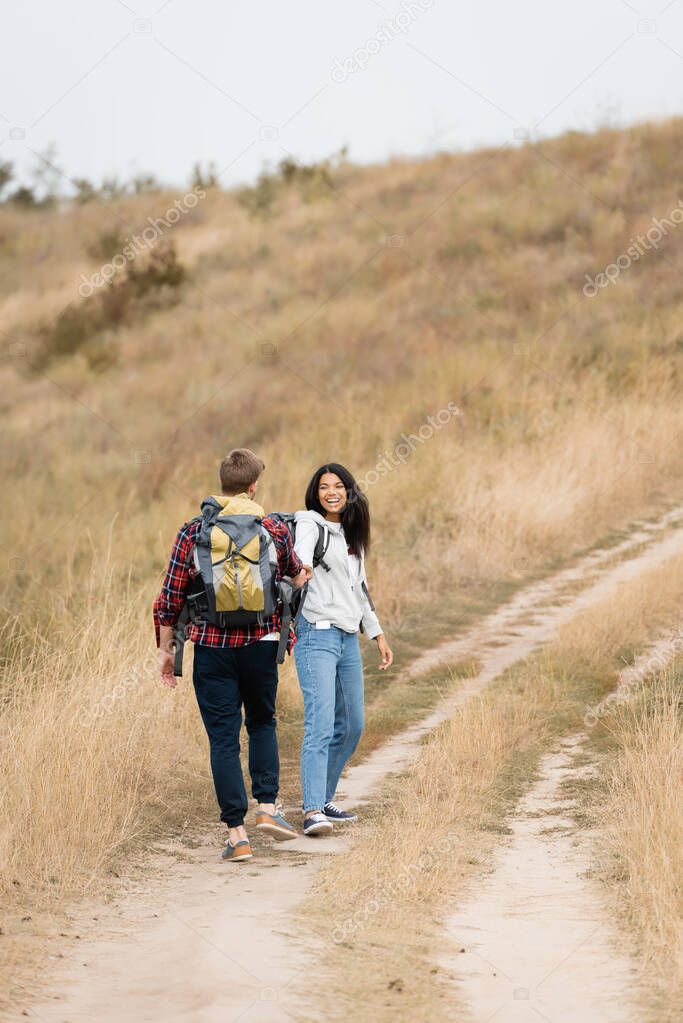 Smiling african american woman looking at boyfriend with backpack while walking on path during trip  