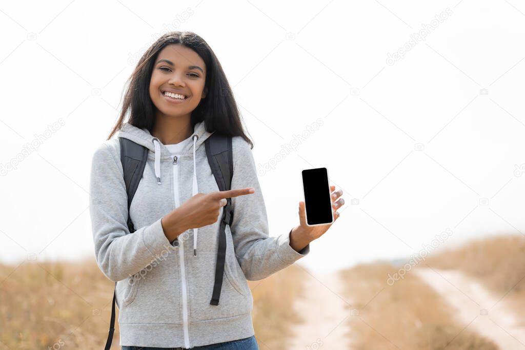 Smiling african american treveller pointing with finger at smartphone with blank screen near path on blurred background 