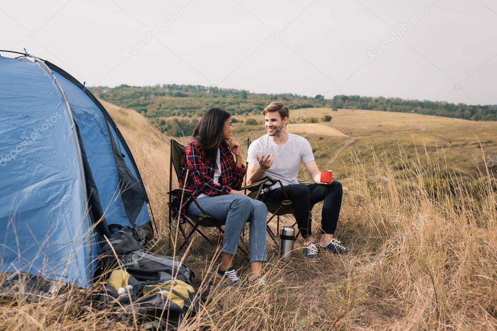 Smiling man with cup talking to african american girlfriend near backpacks and tent on meadow 