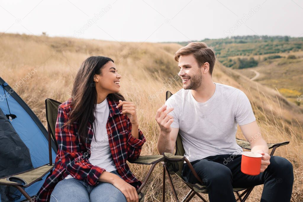 Smiling interracial couple with cup talking on chairs near tent during trip 