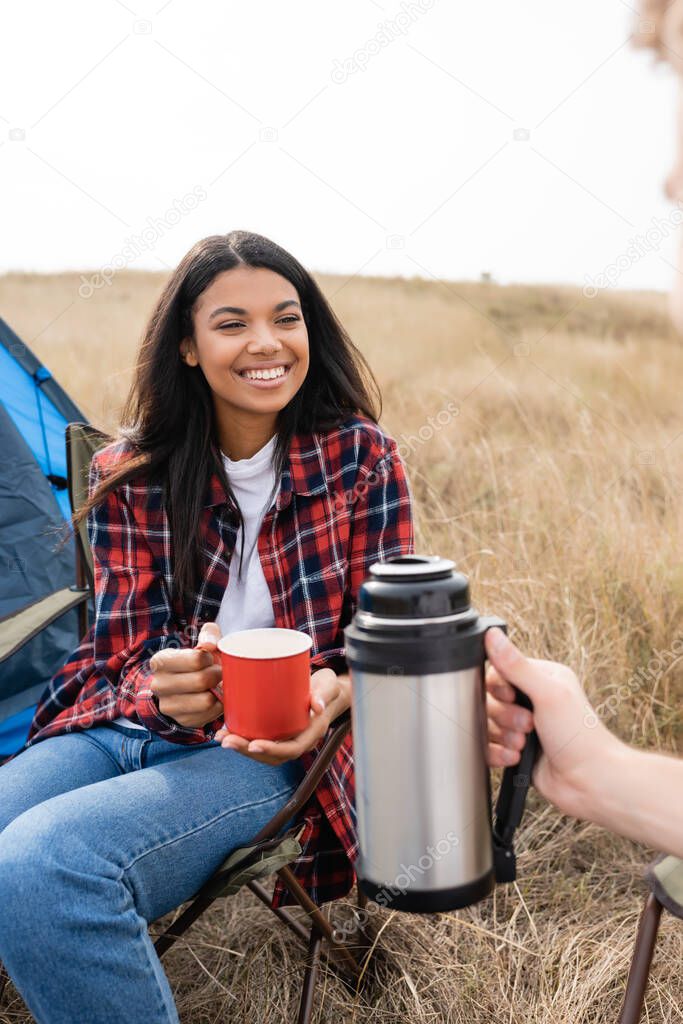 Smiling african american woman holding cup near boyfriend with thermos on blurred foreground and tent on meadow 