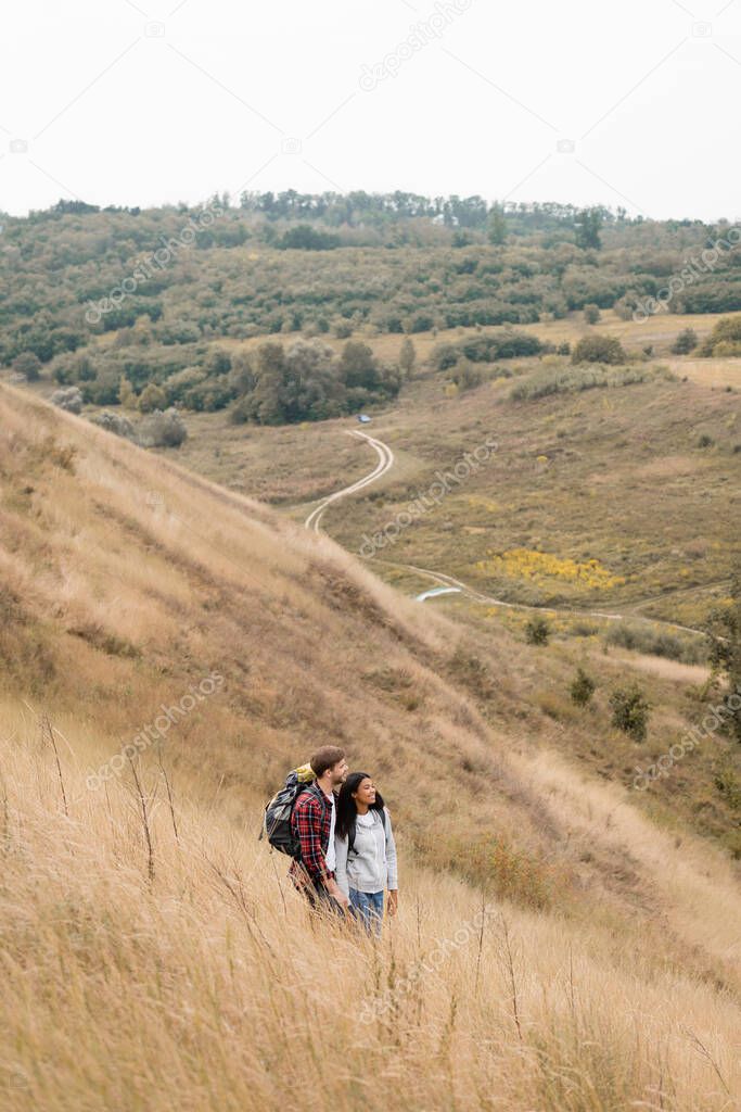 Smiling multiethnic hikers with backpacks looking away on hill with grass on blurred background 