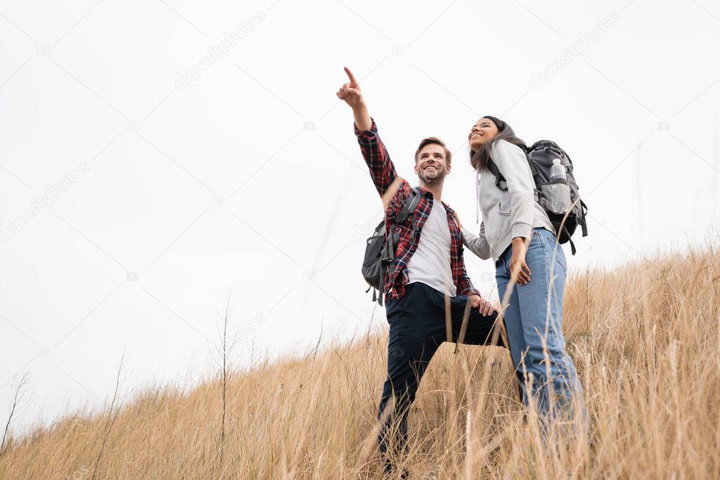 Low angle view of smiling man with backpack pointing with finger away near african american woman while standing on grassy hill with sky at background 