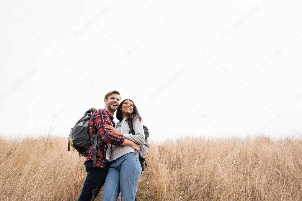 Cheerful multiethnic couple with backpacks hugging on grassy hill with sky at background 