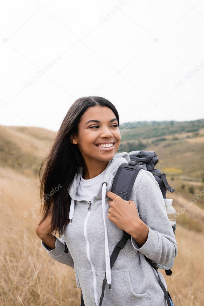 Smiling african american woman touching backpack while standing on hill during trip 