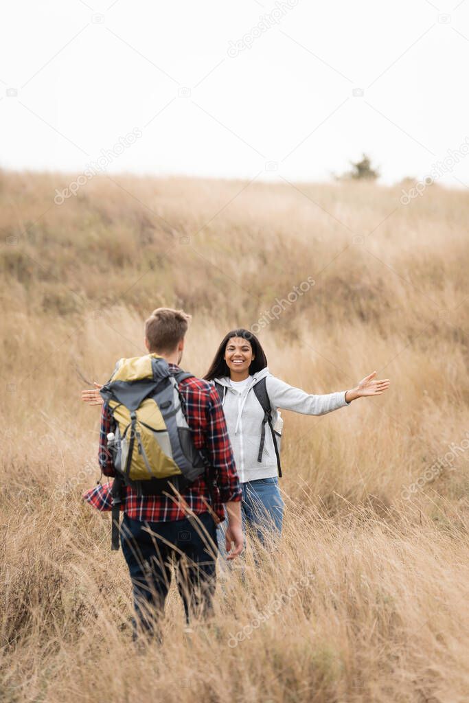 Smiling african american woman smiling at boyfriend with backpack on blurred foreground on grassy hill 