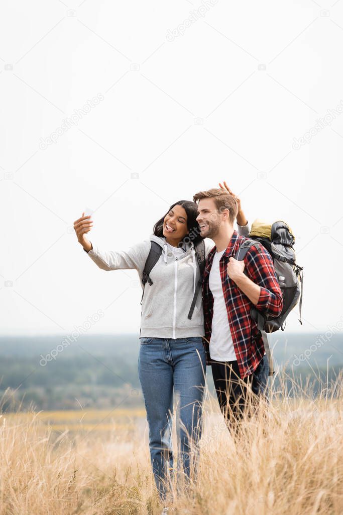 Smiling multiethnic travelers taking selfie with smartphone on meadow 