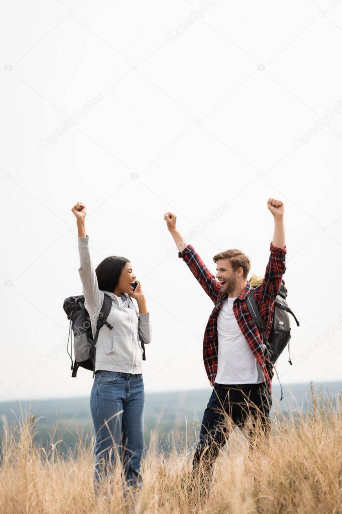 Cheerful multiethnic couple showing yes gesture while talking on smartphone on meadow 