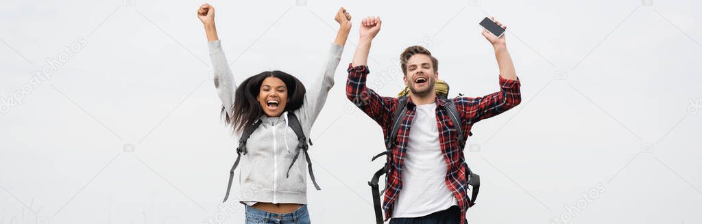 Cheerful multiethnic couple with backpacks and smartphone isolated on grey, banner 