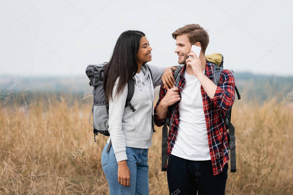 African american woman standing near boyfriend talking on smartphone and touching backpack on lawn 