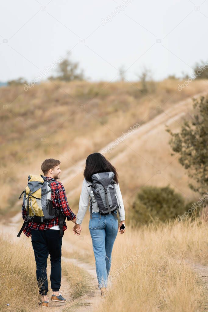 Back view of multiethnic couple with backpacks and smartphone holding hands while walking on path 