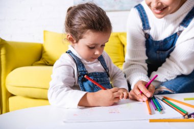 Evdeki annenin yanında kağıt üzerine renkli kalemle çizen kız.